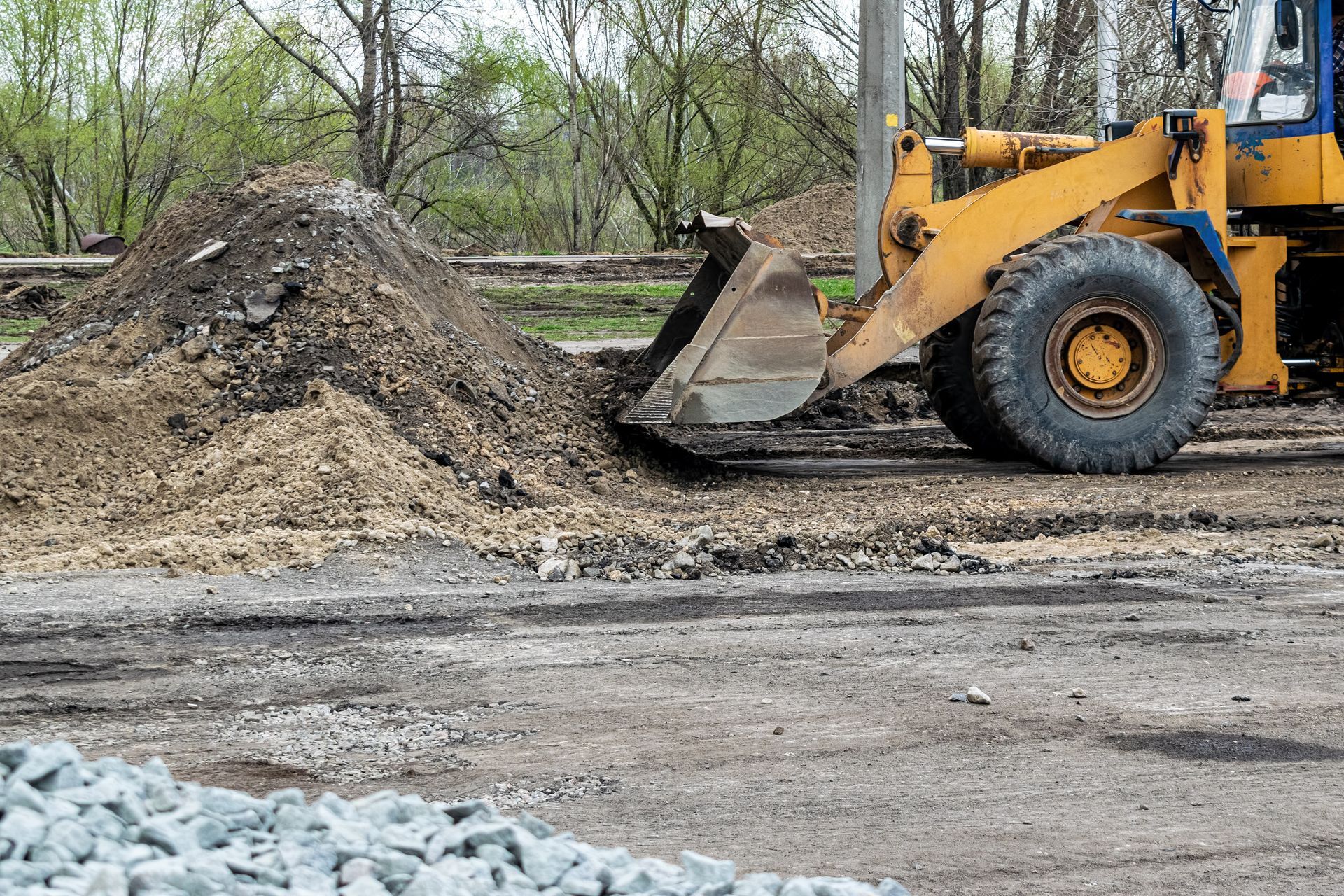 Yellow loader next to a pile of dirt on a gravel road, near a pile of rocks and trees.
