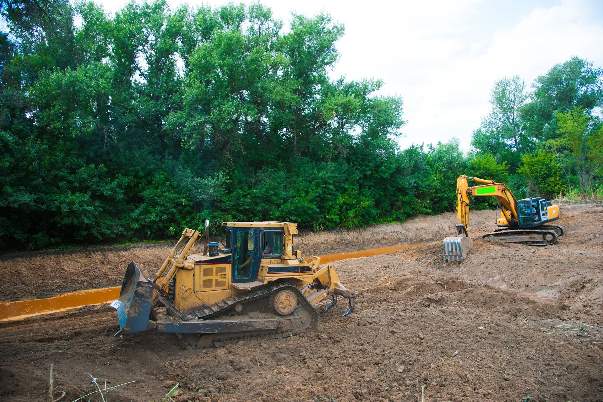 Two construction vehicles on muddy ground clearing land near trees.