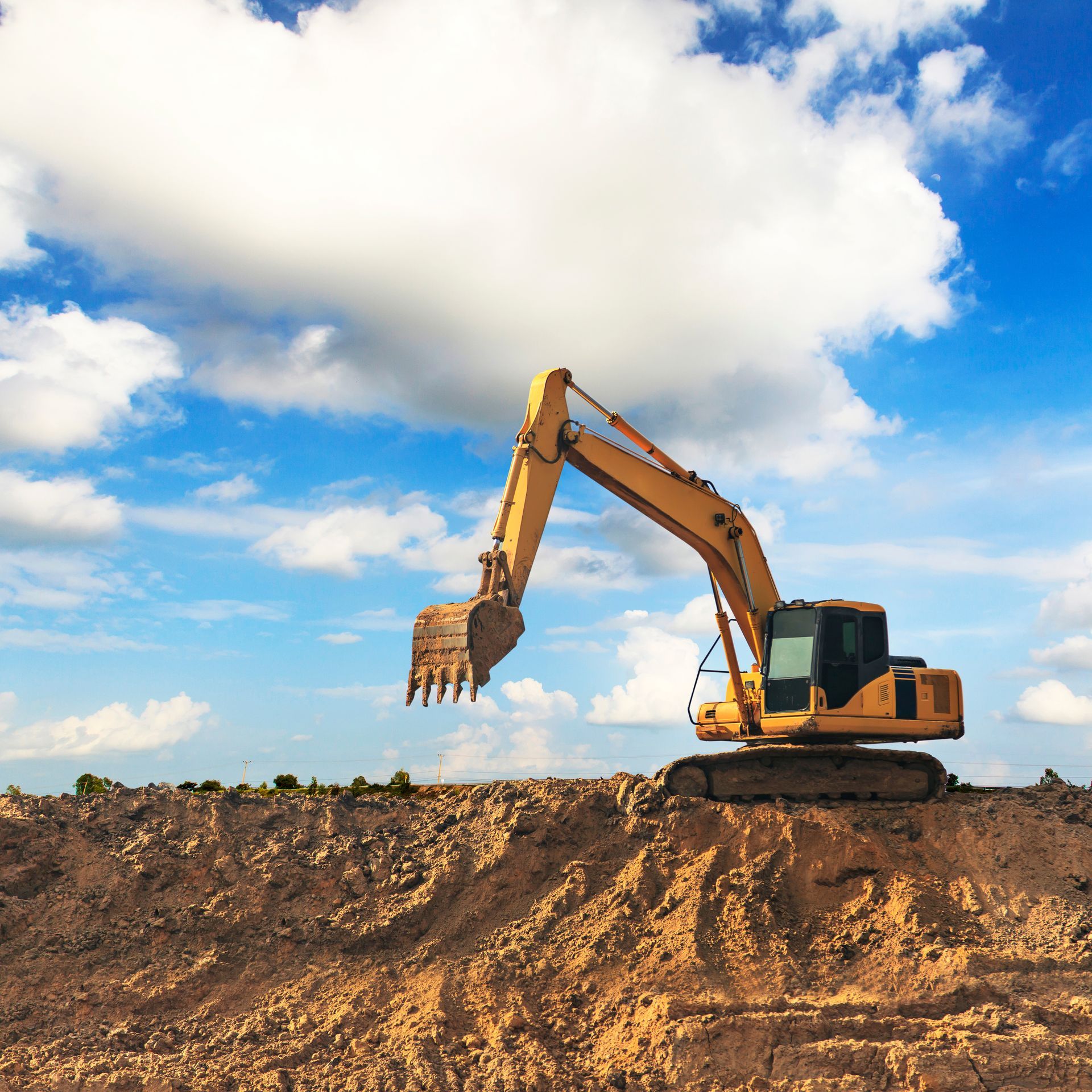 Yellow excavator on a dirt hill against a blue sky with fluffy white clouds.