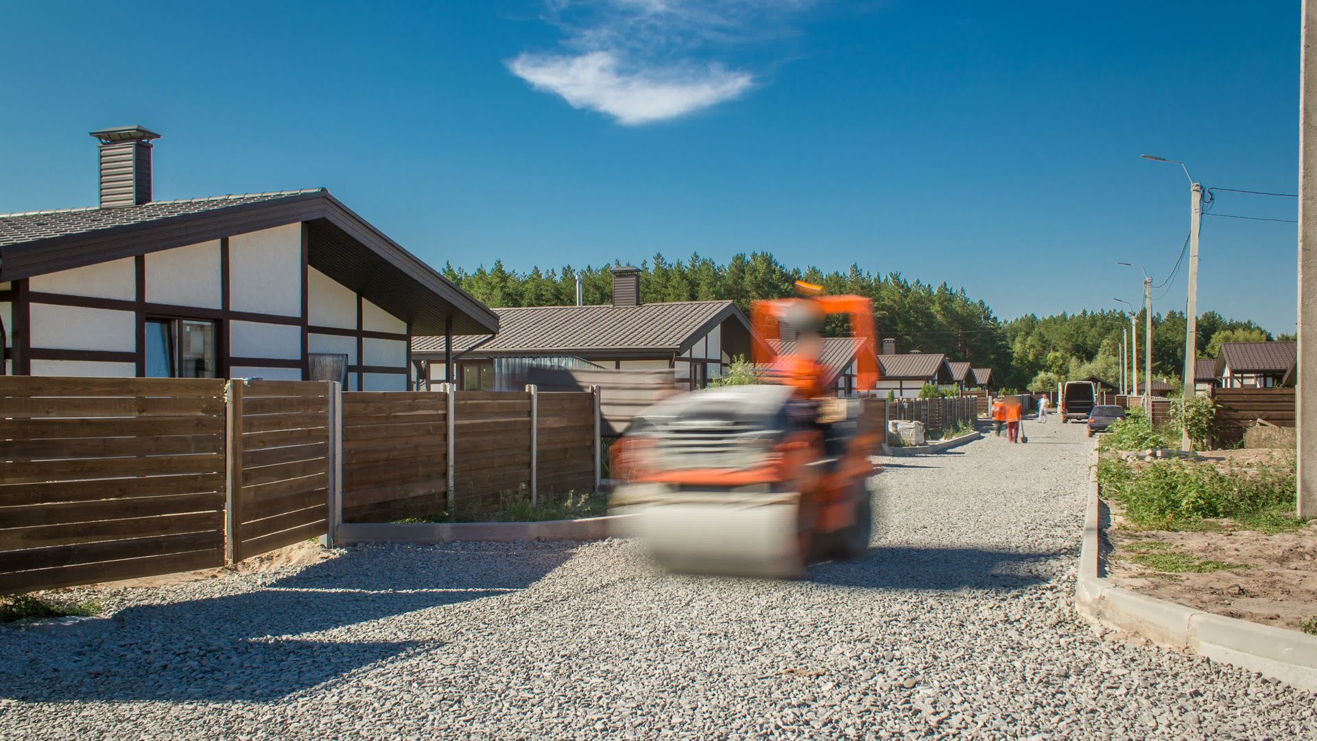 Road roller compacting gravel road in a residential area, under a blue sky.
