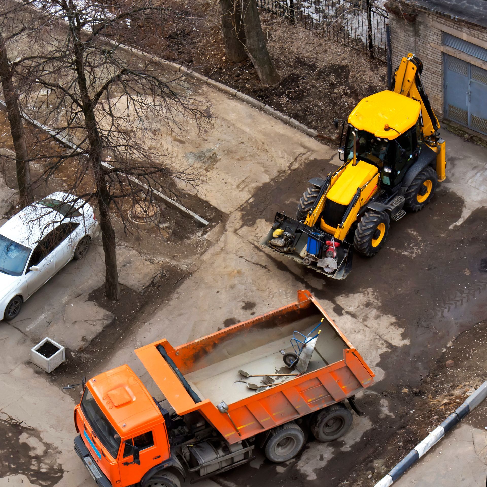 Yellow backhoe loading an orange dump truck on a concrete surface next to a white car.