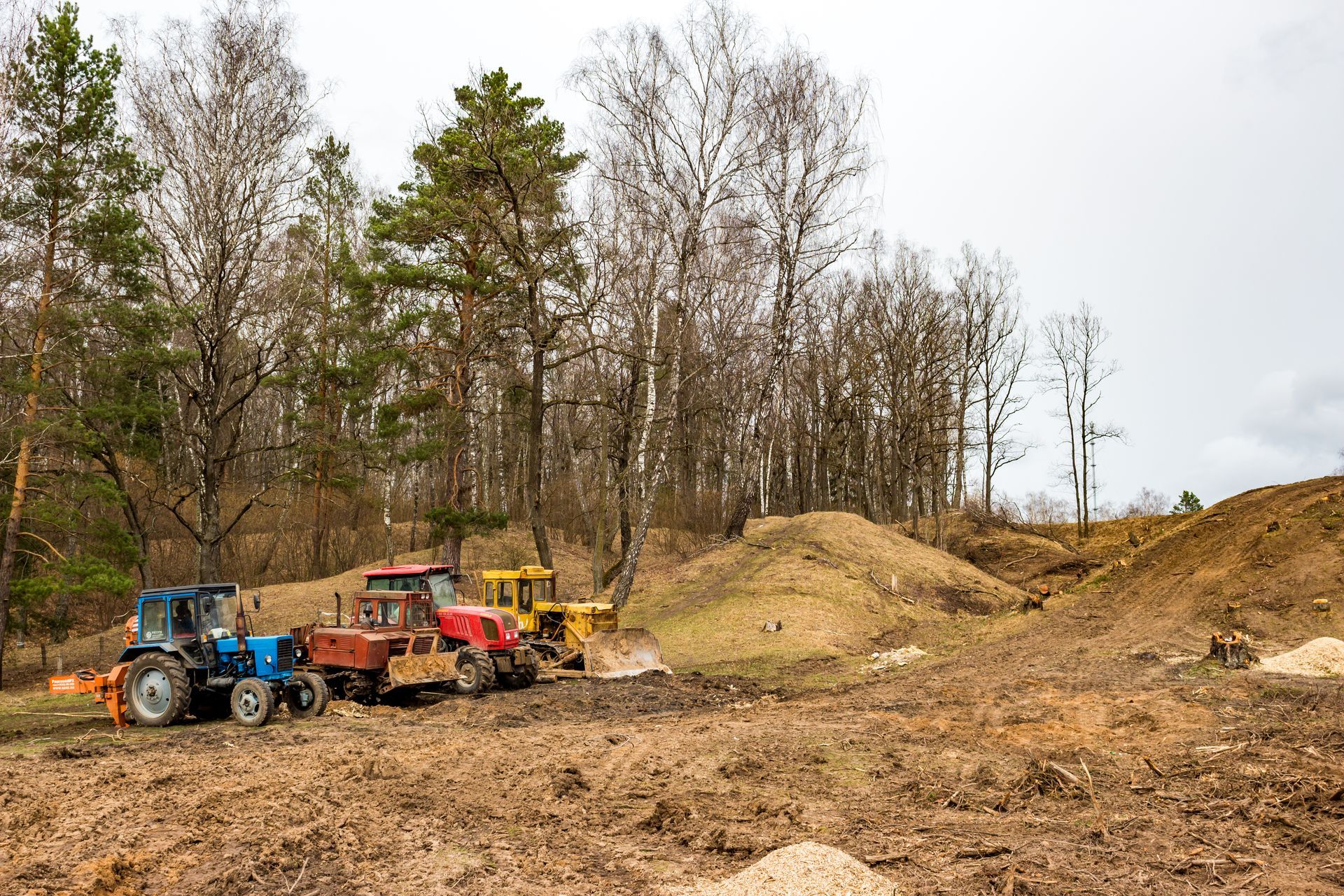 Tractors lined up in a muddy clearing, preparing to dig. Brown soil, trees in background.