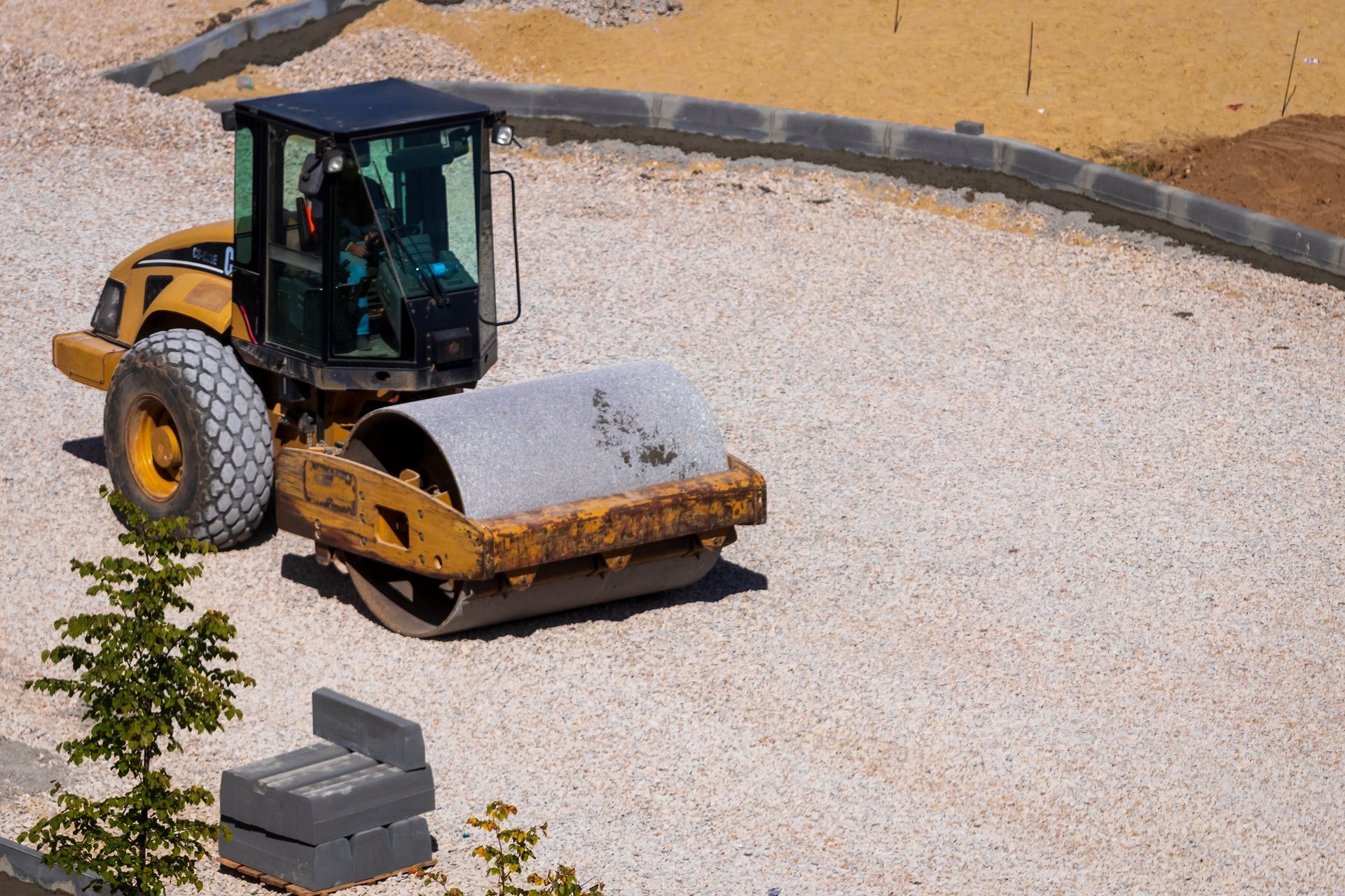 Road roller compacting gravel surface. A person is driving the machine near a curb.