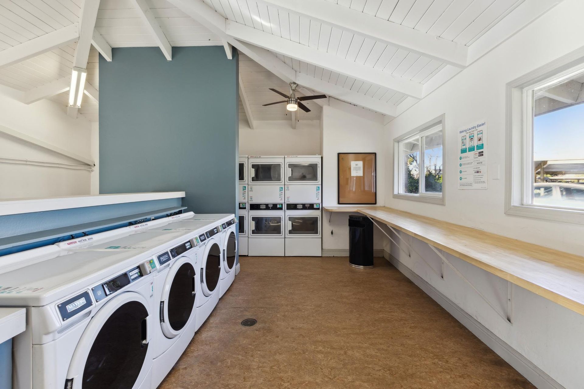Communal laundry room with front-loading washers and dryers, a long wooden counter, and high vaulted white ceilings.