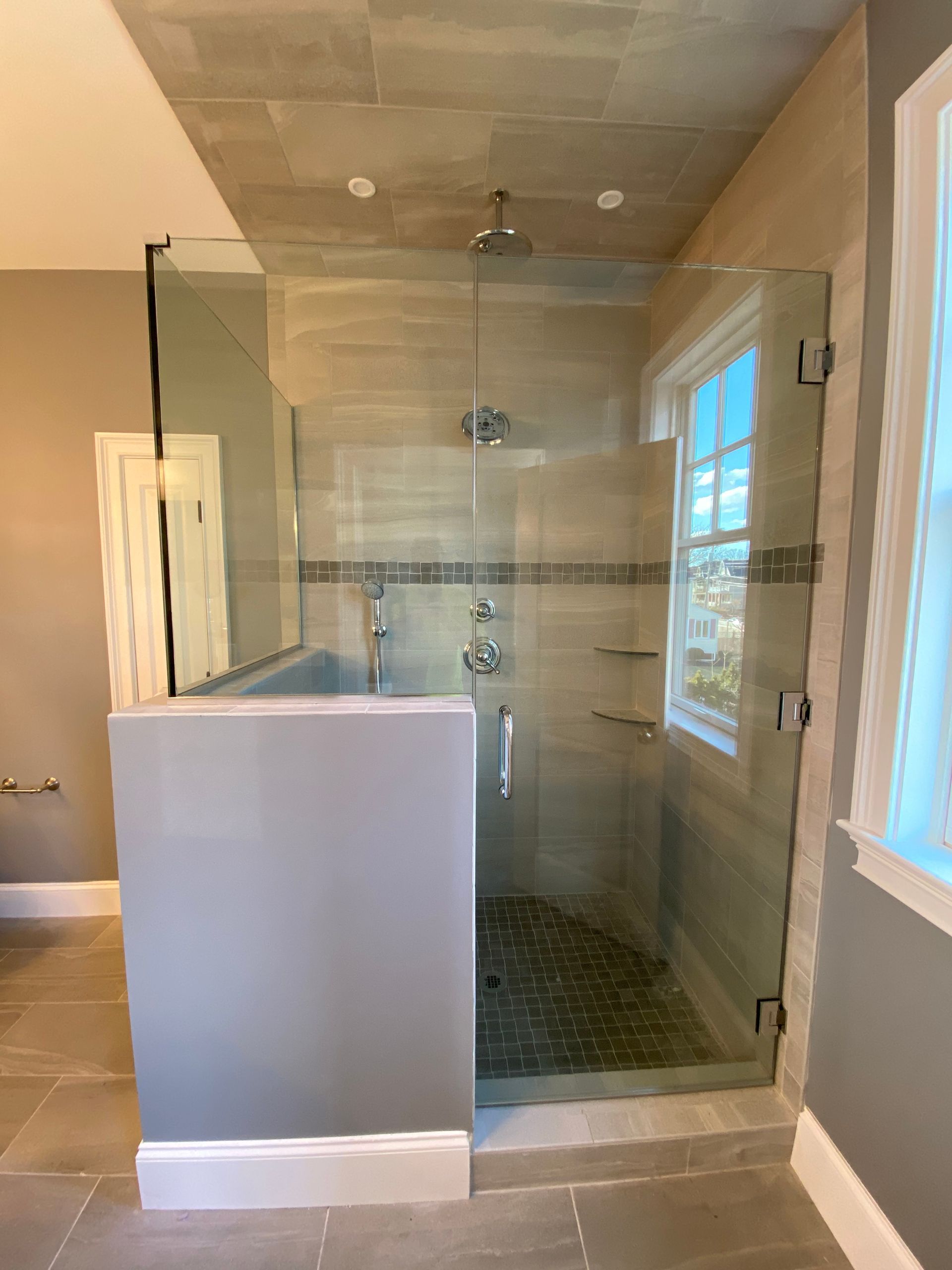 Glass-walled shower with beige tile, a window, and a gray partition. The shower head and controls are visible.