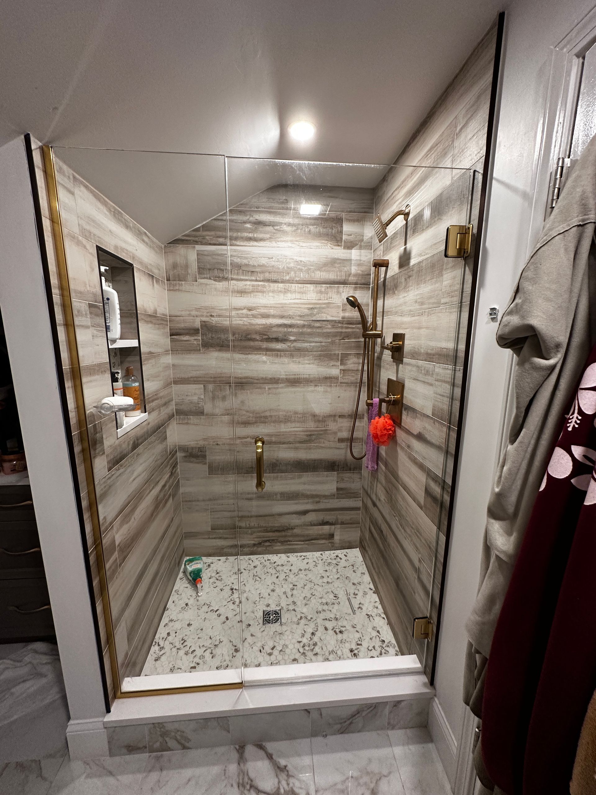 Shower with gold fixtures, wood-look tile, and mosaic floor.  Clear glass door.  Bathroom setting.
