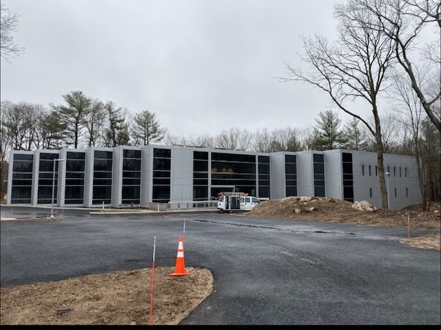 Gray modern building under construction with large windows, bare trees, and overcast sky.