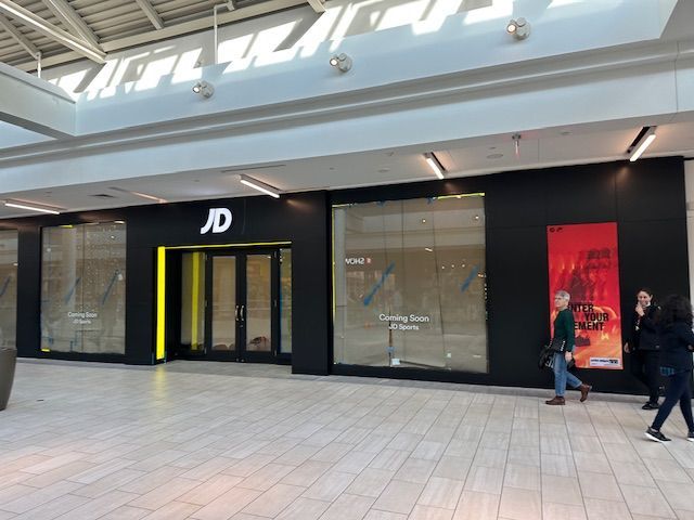JD Sports store under construction in a mall. Black exterior with yellow accents, two people walking past.