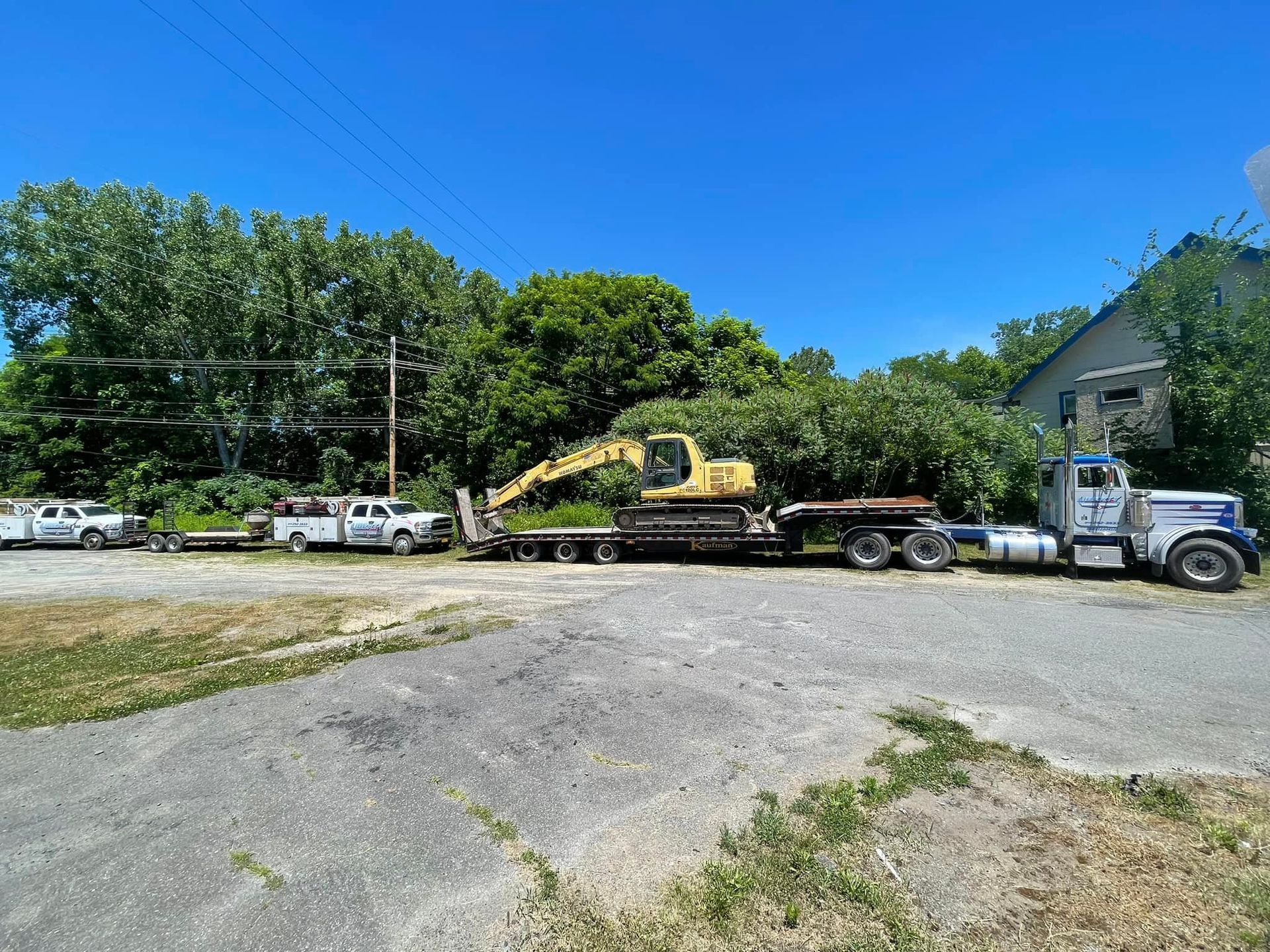 A flatbed truck carries a yellow excavator in a gravel lot next to two white service trucks under a clear blue sky.
