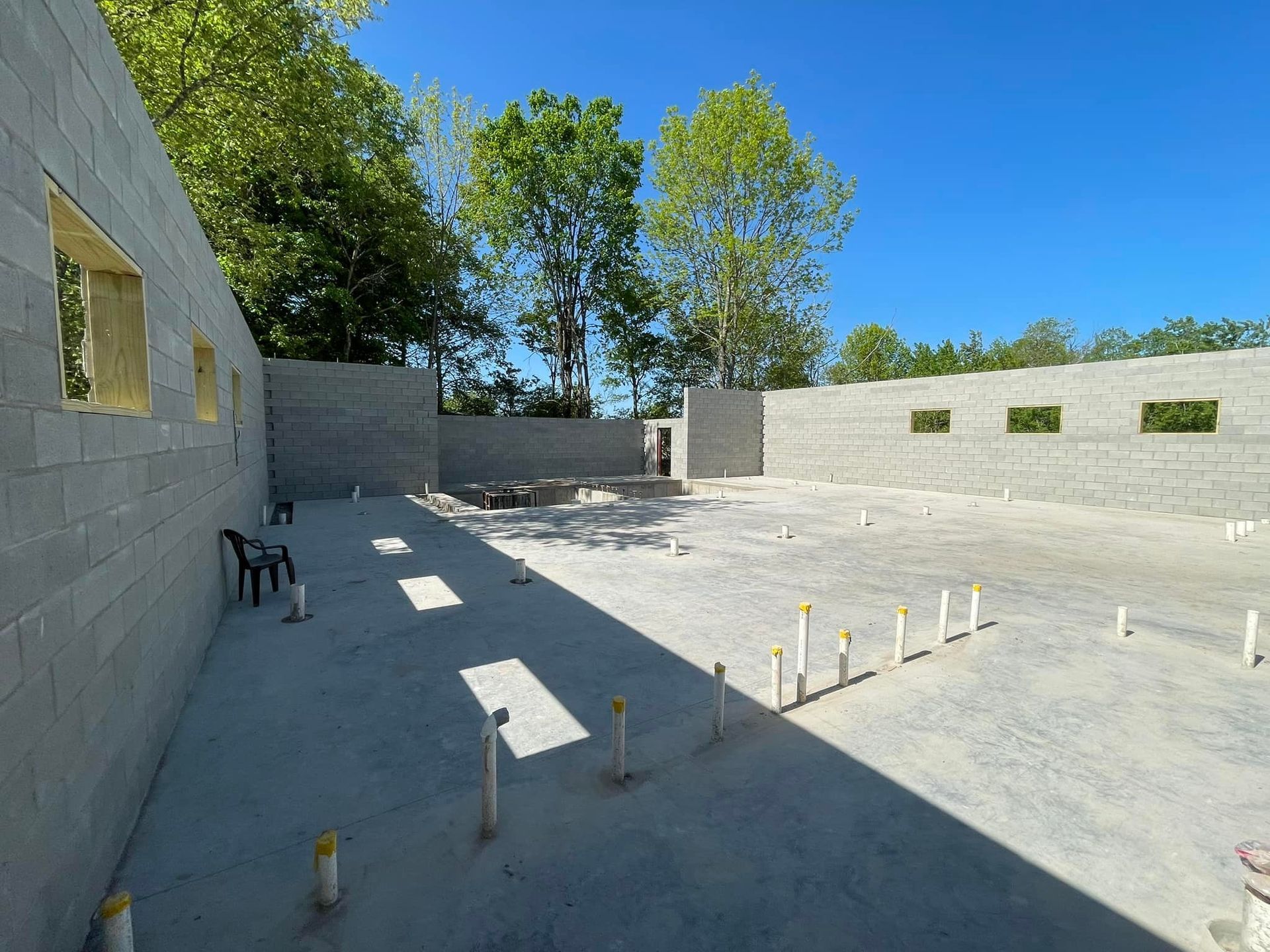 Construction site with grey concrete block walls, a concrete floor with plumbing pipes, and trees against a blue sky.