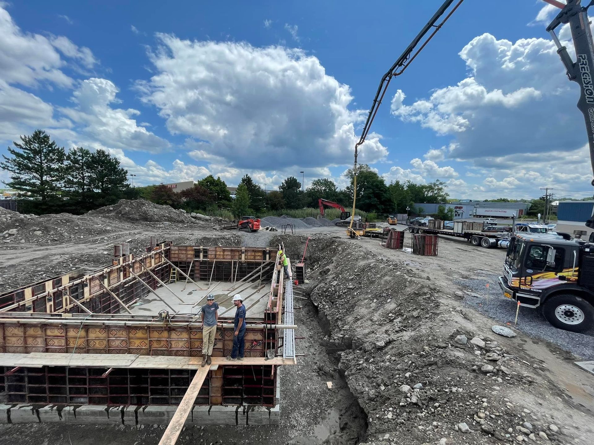 Two people stand on a wood platform overlooking a rectangular concrete foundation under construction on a sunny day.