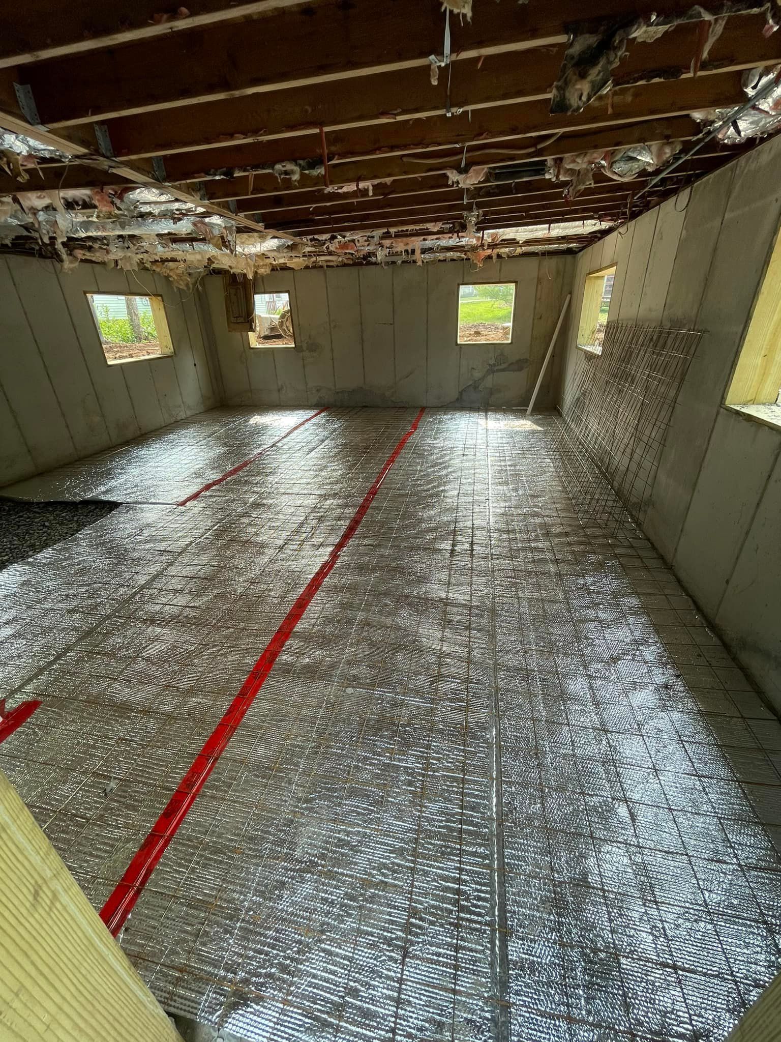 An unfinished basement interior with concrete walls, exposed ceiling joists, and a reflective silver underlayment floor.