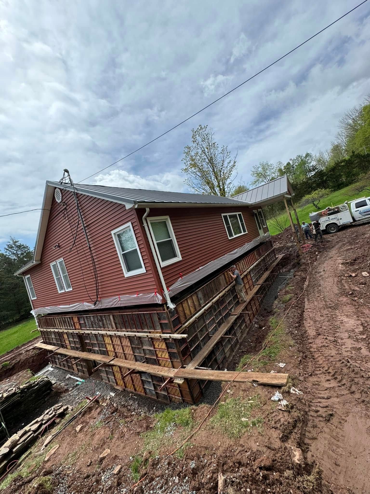 A small red house with a metal roof is elevated on wooden support beams above a muddy construction site.