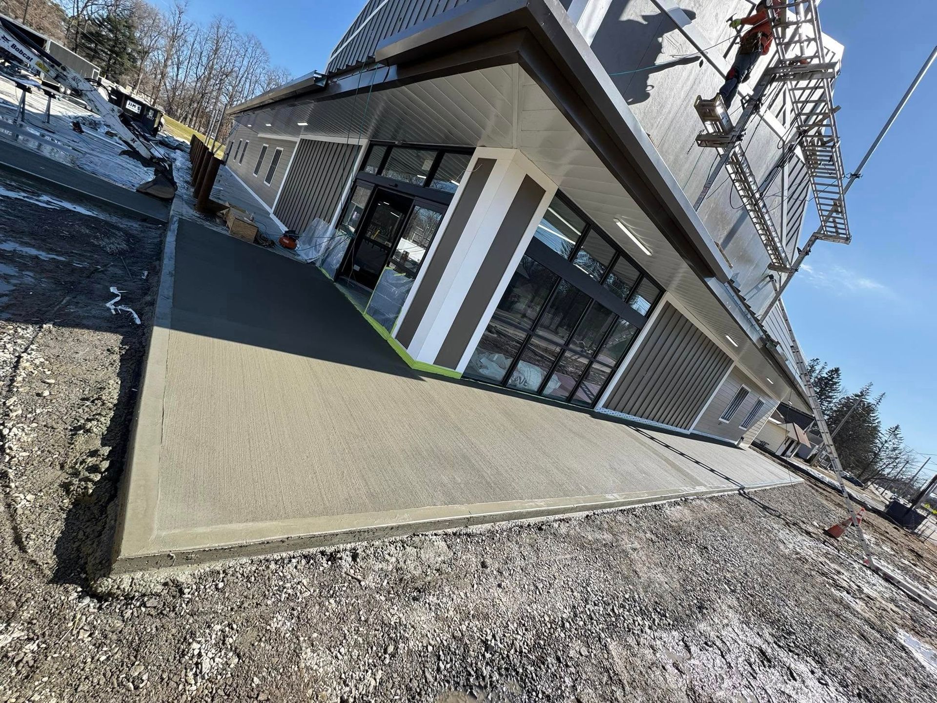 A newly poured concrete walkway wraps around the corner of a modern retail building under a clear, sunny sky.