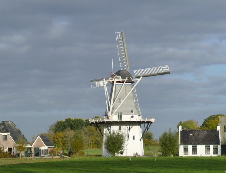 Een witte windmolen met draaiende wieken, in een groen veld, onder een bewolkte hemel.
