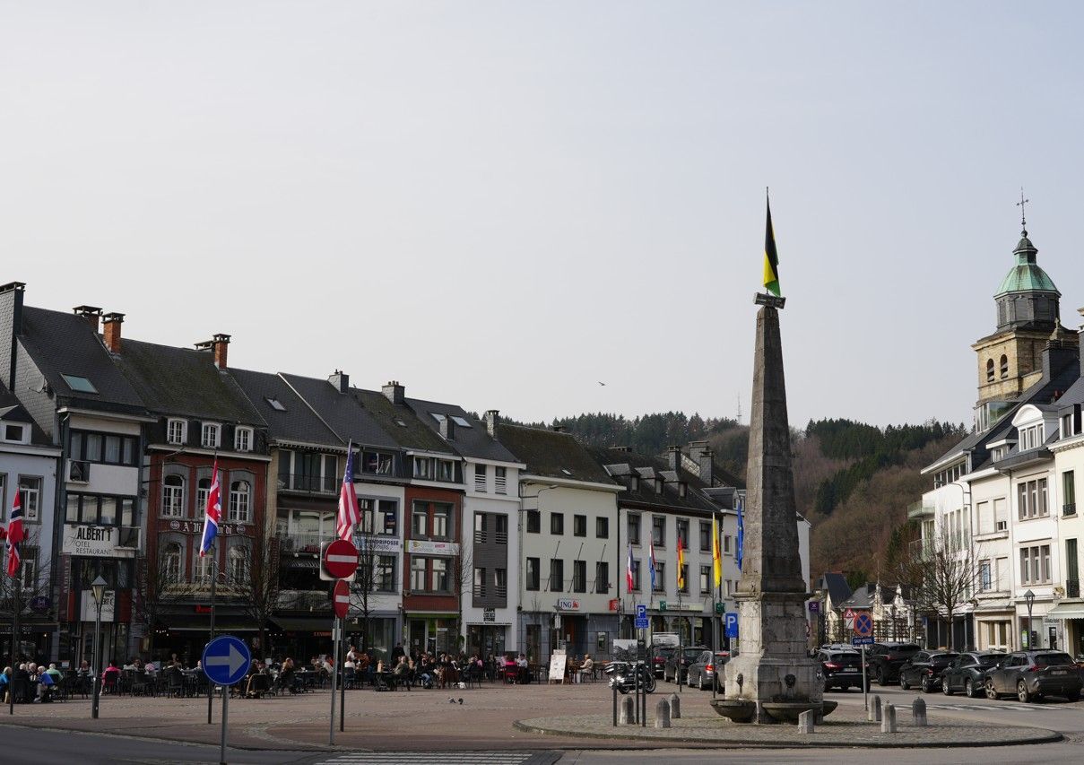 Een historisch stadsplein met flatgebouwen, terrasjes, een obelisk en vlaggen onder een heldere hemel.