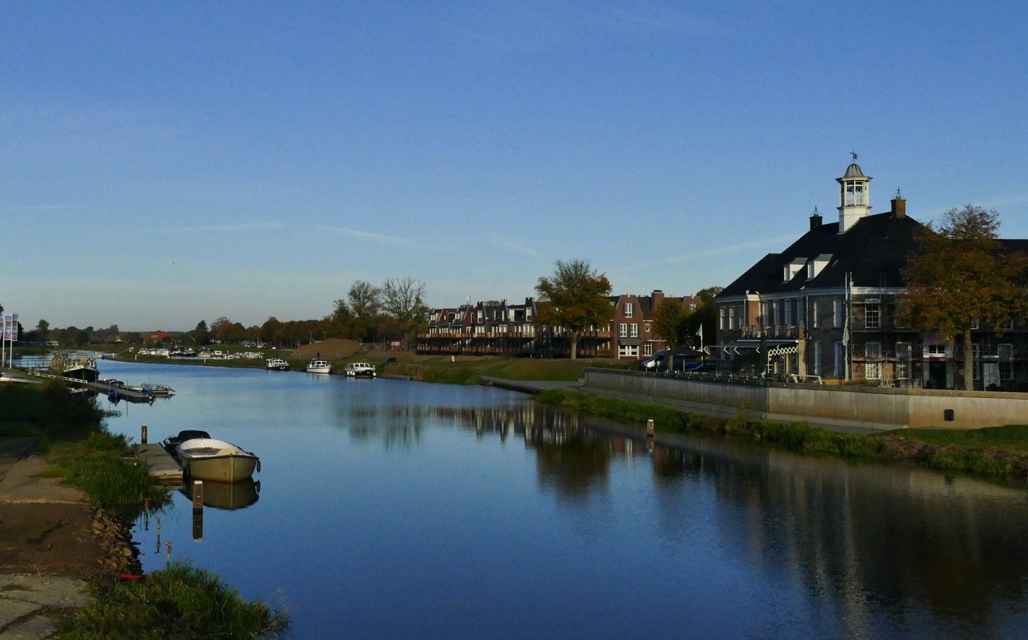 Een rustige rivier stroomt langs een traditioneel gebouw met een klokkentoren en met bomen. Duidelijk Ommen