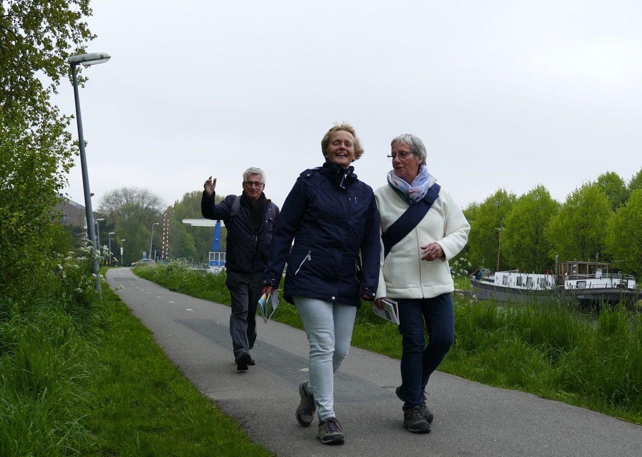 Drie mensen lopen over een pad langs een waterweg. Een van hen zwaait. Bewolkte lucht, bomen.