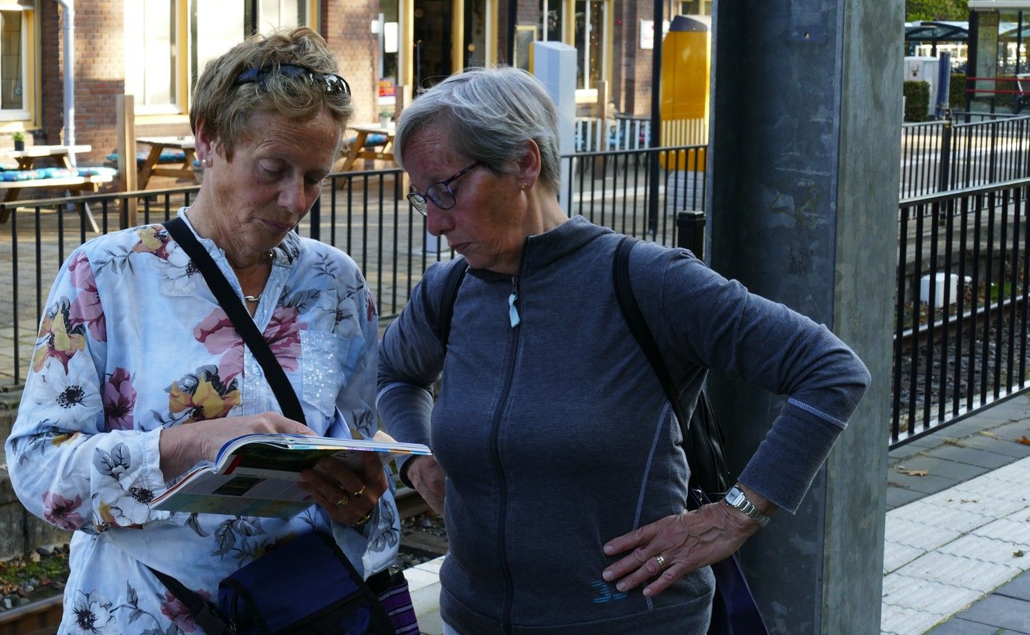 Twee mensen staan ​​buiten bij een metalen hek en kijken samen naar een open brochure of kaart.