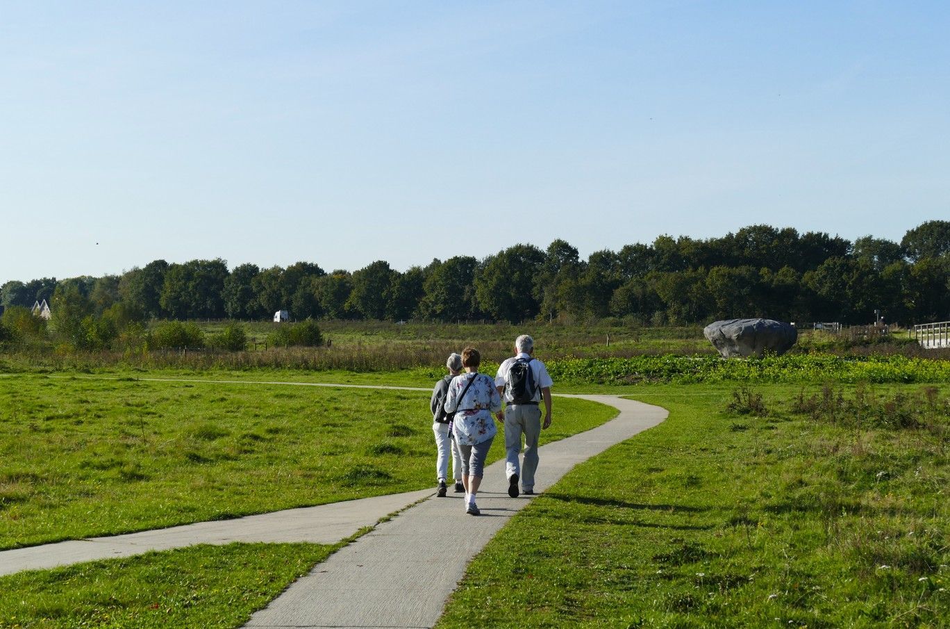 Drie mensen lopen over een geplaveid pad door een grasveld richting een rij bomen onder een helderblauwe hemel.