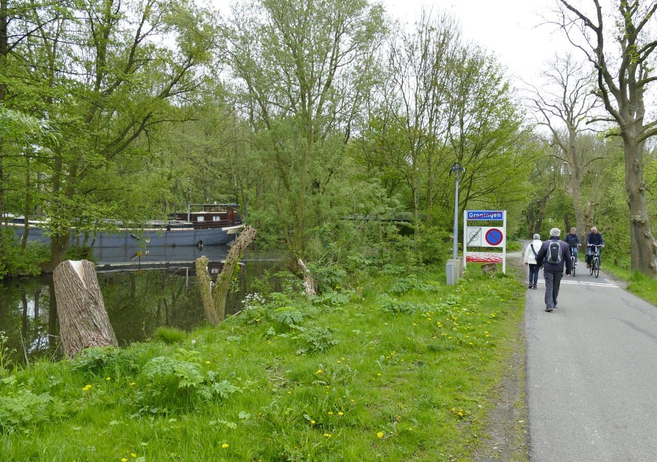Pad langs een kanaal, met een bord en mensen die erlangs lopen; bomen en een boot zijn zichtbaar.