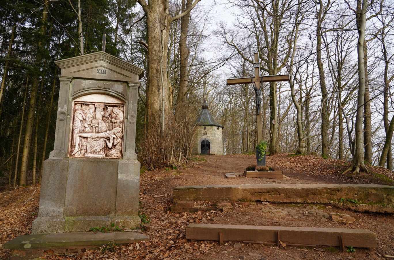 Op een bebost pad op een heuveltop, dat naar een kleine, ronde kapel leidt, staan een stenen schrijn en een groot houten kruis.