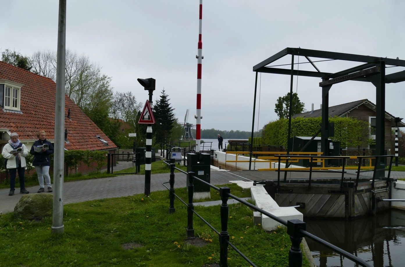 Een verhoogde brug met een rood-wit gestreepte balustrade; mensen staan ​​er vlakbij, op de achtergrond zijn een kanaal en huizen te zien.