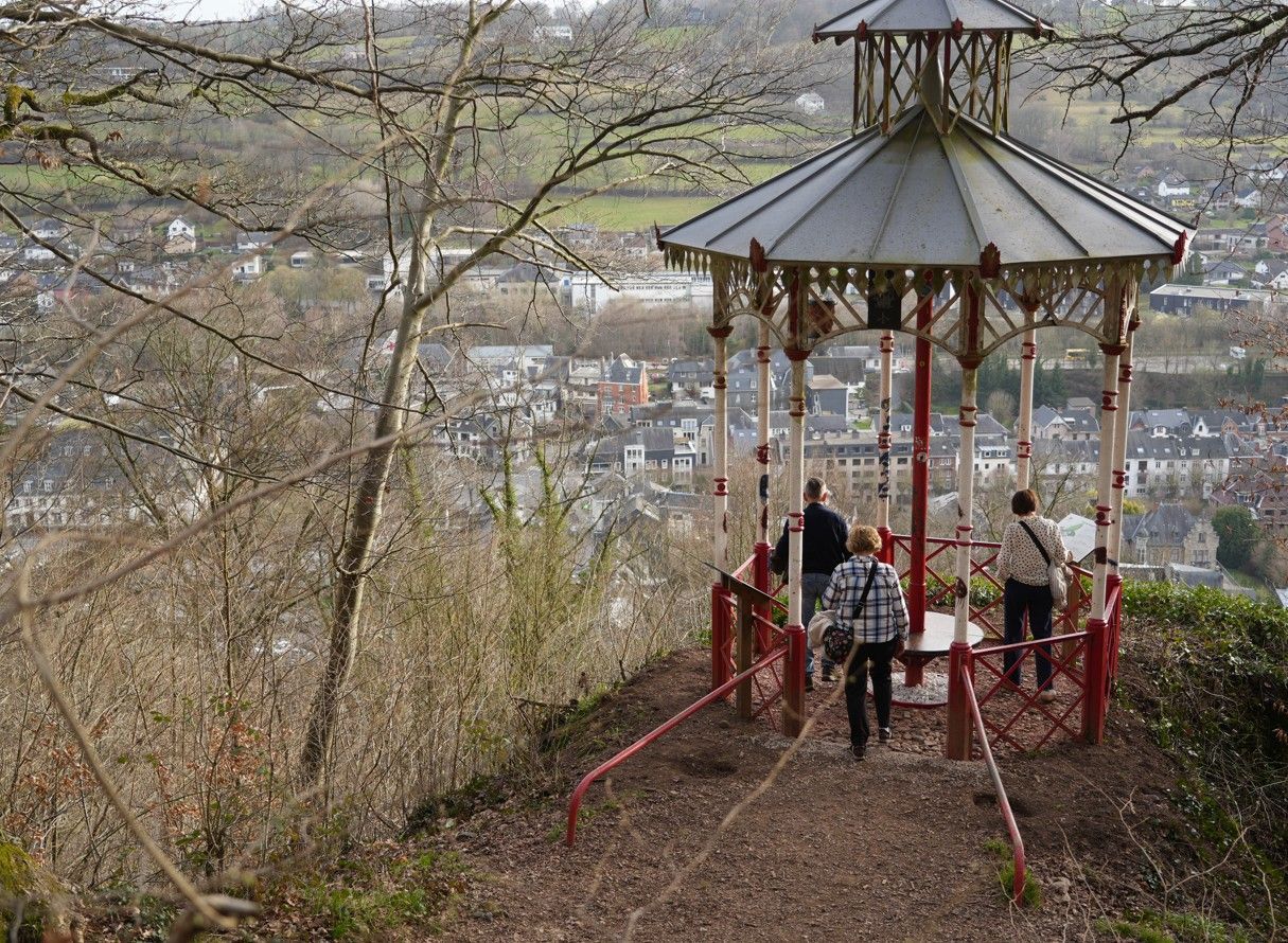 Drie mensen staan in een wit-rood prieel met uitzicht op een stadje in een bosrijk, heuvelachtig landschap.