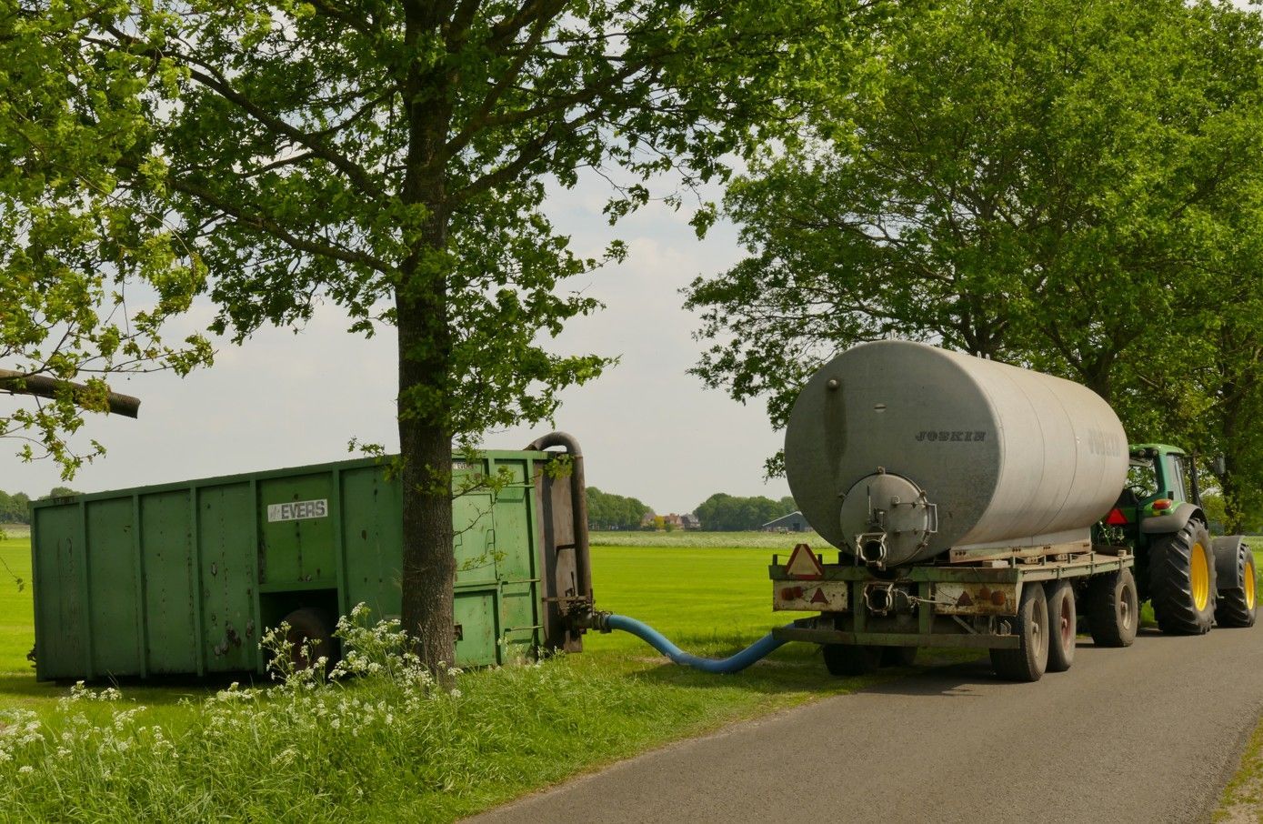 Een tractor trekt een tankwagen die vloeistof in een groene container pompt, vlakbij een boom op een landweg.