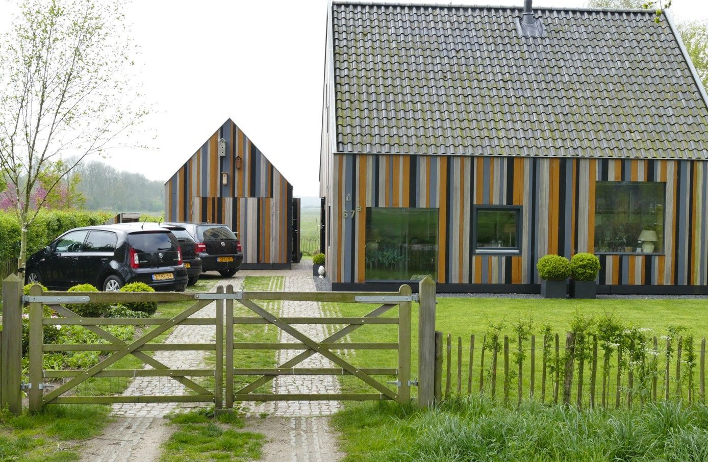 Huis met verticale houten gevelbekleding, oprit en auto's. Poort en kleine garage op de achtergrond.