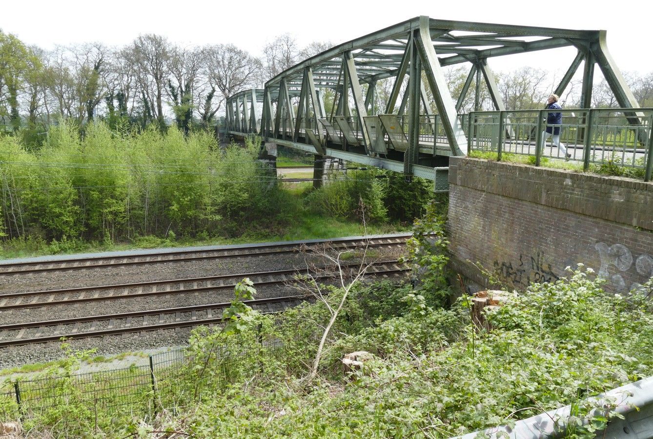 Groene metalen brug over spoorrails en groen, persoon loopt over de brug.