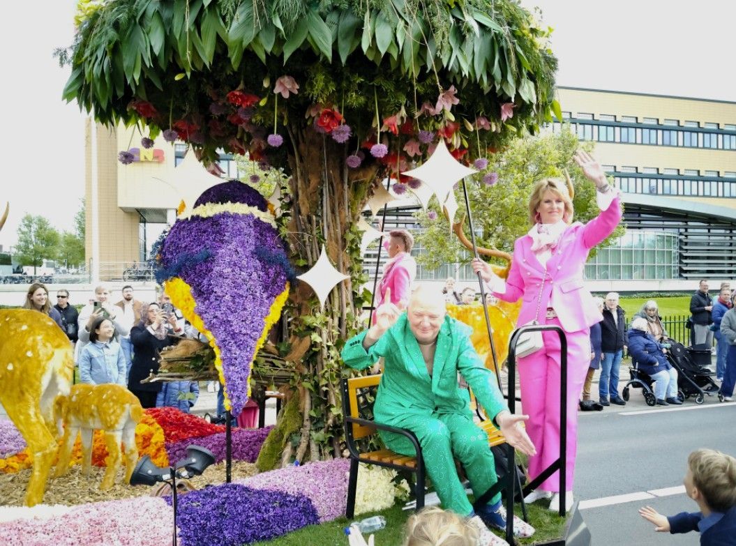 Een kleurrijke praalwagen met paarse bloemen en twee artiesten in kostuums, waarvan er één zit en de ander zwaait.
