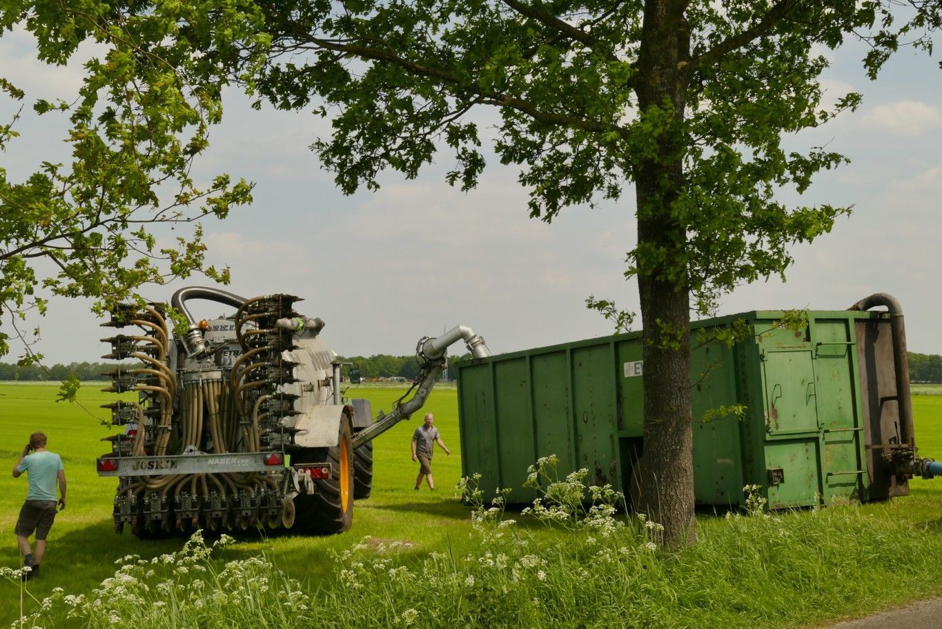 Landbouwmachine naast een groene container op een grasveld met mensen in de buurt.