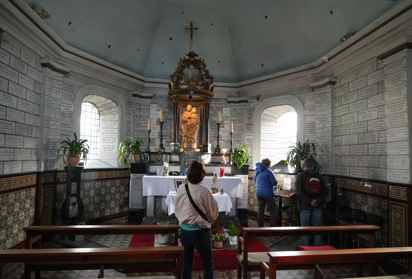 Een blik vanaf de achterkant van een kleine kapel, met drie mensen die voor een altaar staan dat versierd is met gouden ornamenten.