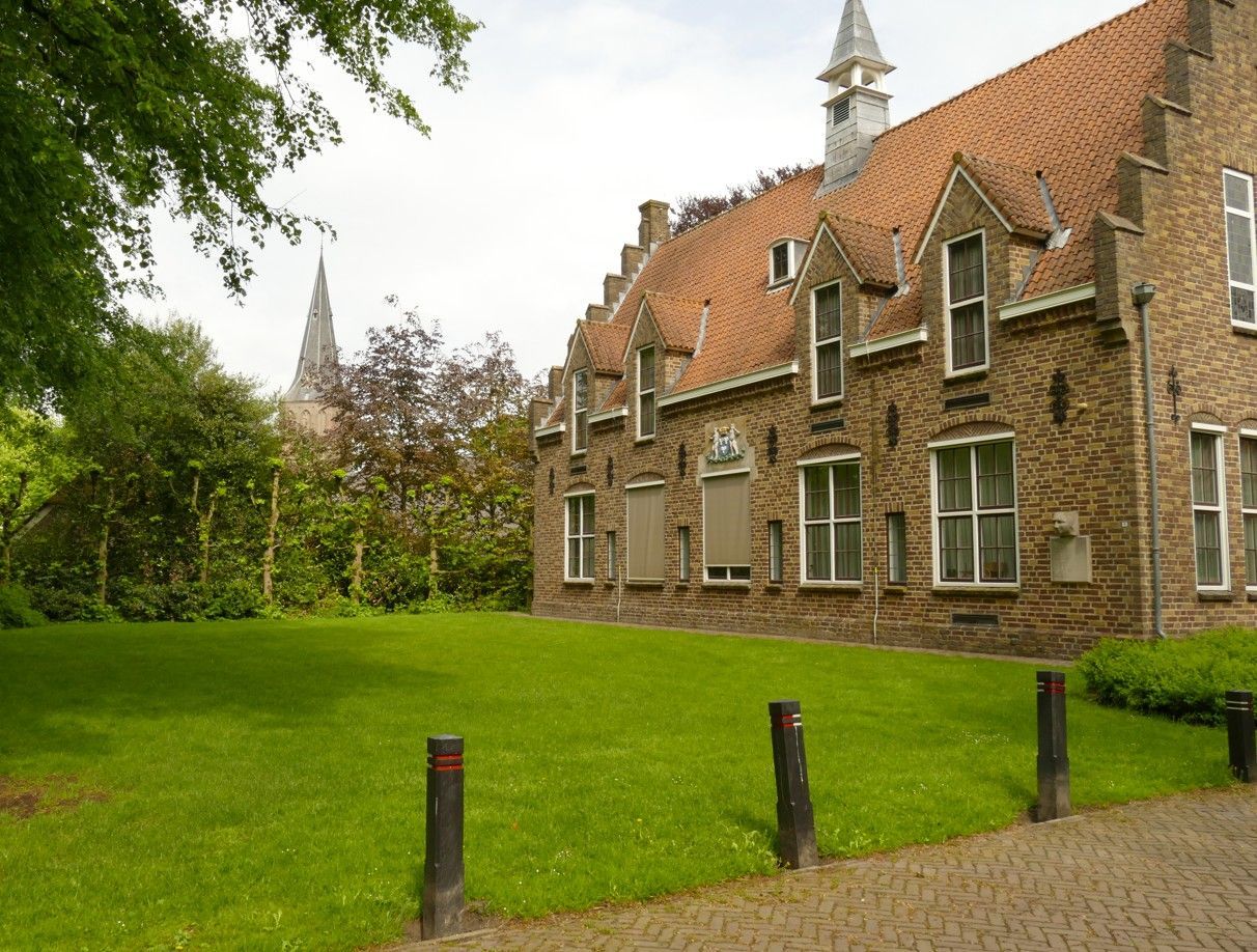 Een bakstenen gebouw met een rood pannendak en een groen gazon, met een kerktoren op de achtergrond.