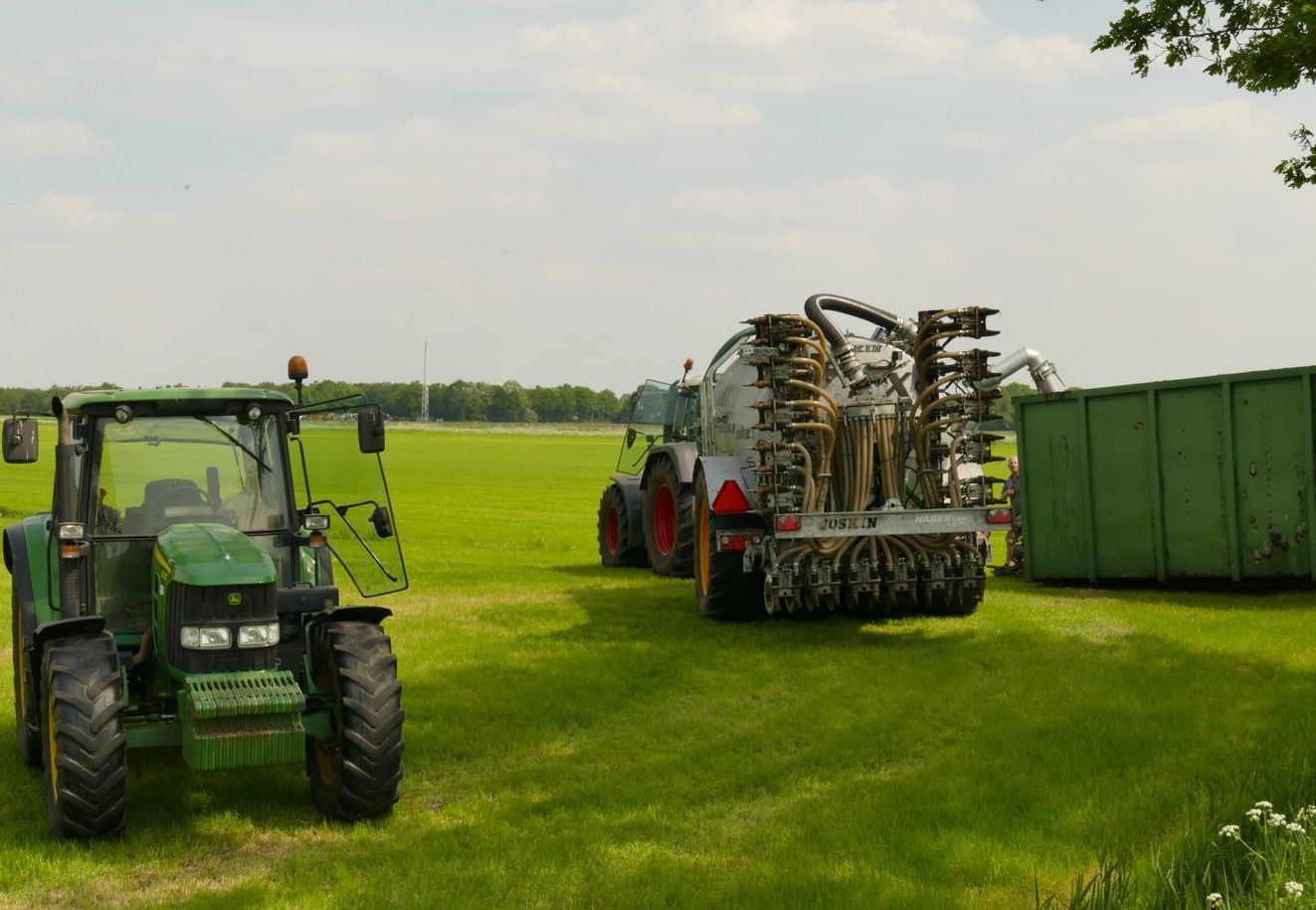 Groene tractor en tankwagen die materiaal verspreiden op een grasveld. Op de achtergrond is een groene container te zien.