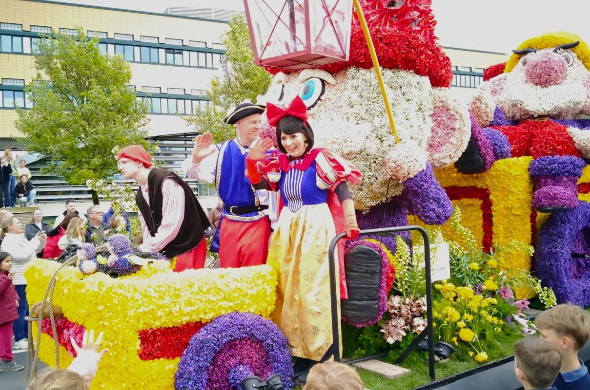Kleurrijke praalwagen met mensen in kostuums, bloemen en een kasteelachtige versiering buiten.