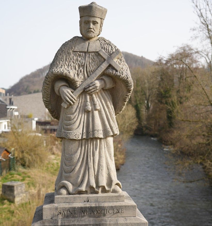 Een stenen beeld van een religieuze figuur die een kruis vasthoudt, staande op een brug over een rivier met een heuvel op de achtergrond.