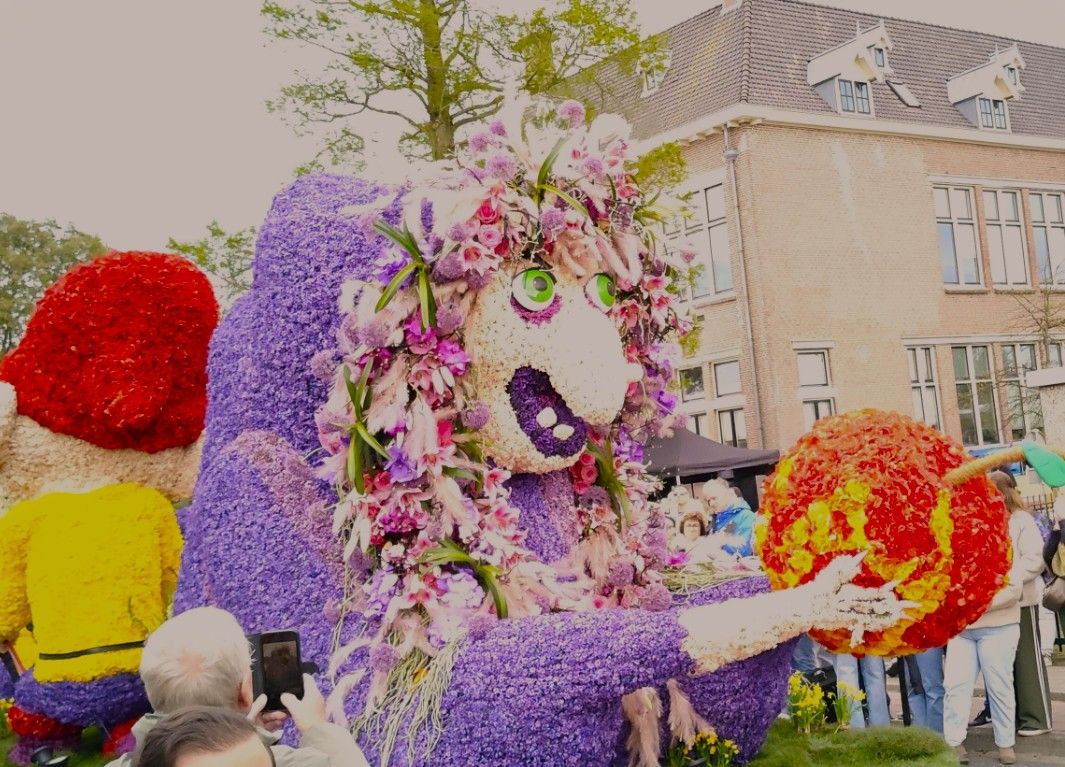 Een kleurrijke praalwagen met bloemen, een grote paarse dierenfiguur en rood-gele kostuums rijdt door een stadsstraat.