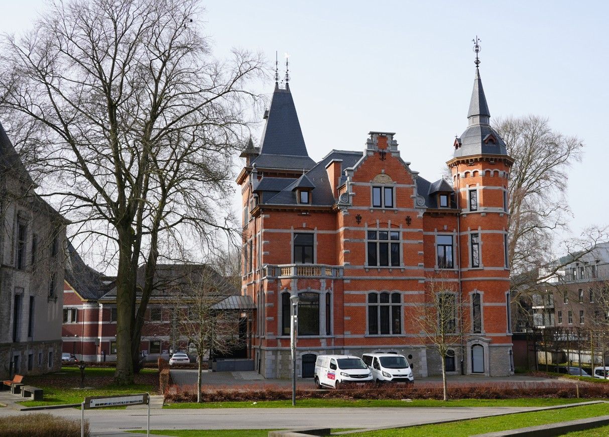Een herenhuis van meerdere verdiepingen, opgetrokken uit rode baksteen, met een torentje, een steil leien dak en witte stenen accenten, gelegen tussen de bomen op een zonnige dag.