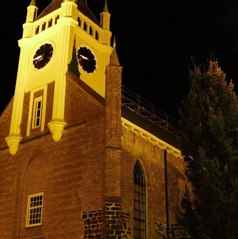 De kerk in Ommen die 's nachts verlicht is, gezien van onderaf tegen een donkere hemel naast een boom.
