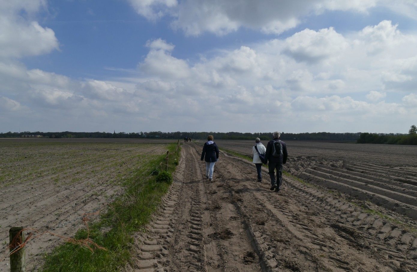 Drie mensen lopen over een modderig pad door een veld onder een bewolkte hemel.