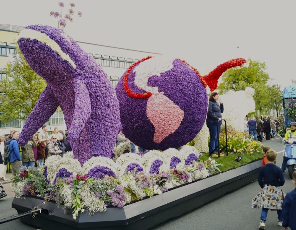 Paarse, met bloemen versierde praalwagen in de vorm van een walvis met rode accenten, omringd door bloemen.