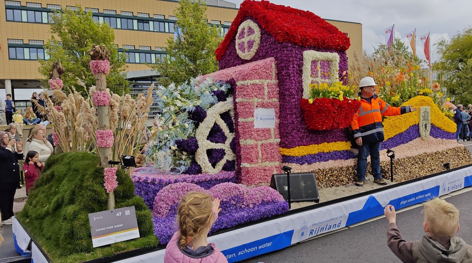 Een kleurrijke praalwagen met bloemen, een groot roze huis, een arbeider in oranje en toeschouwers langs de route.