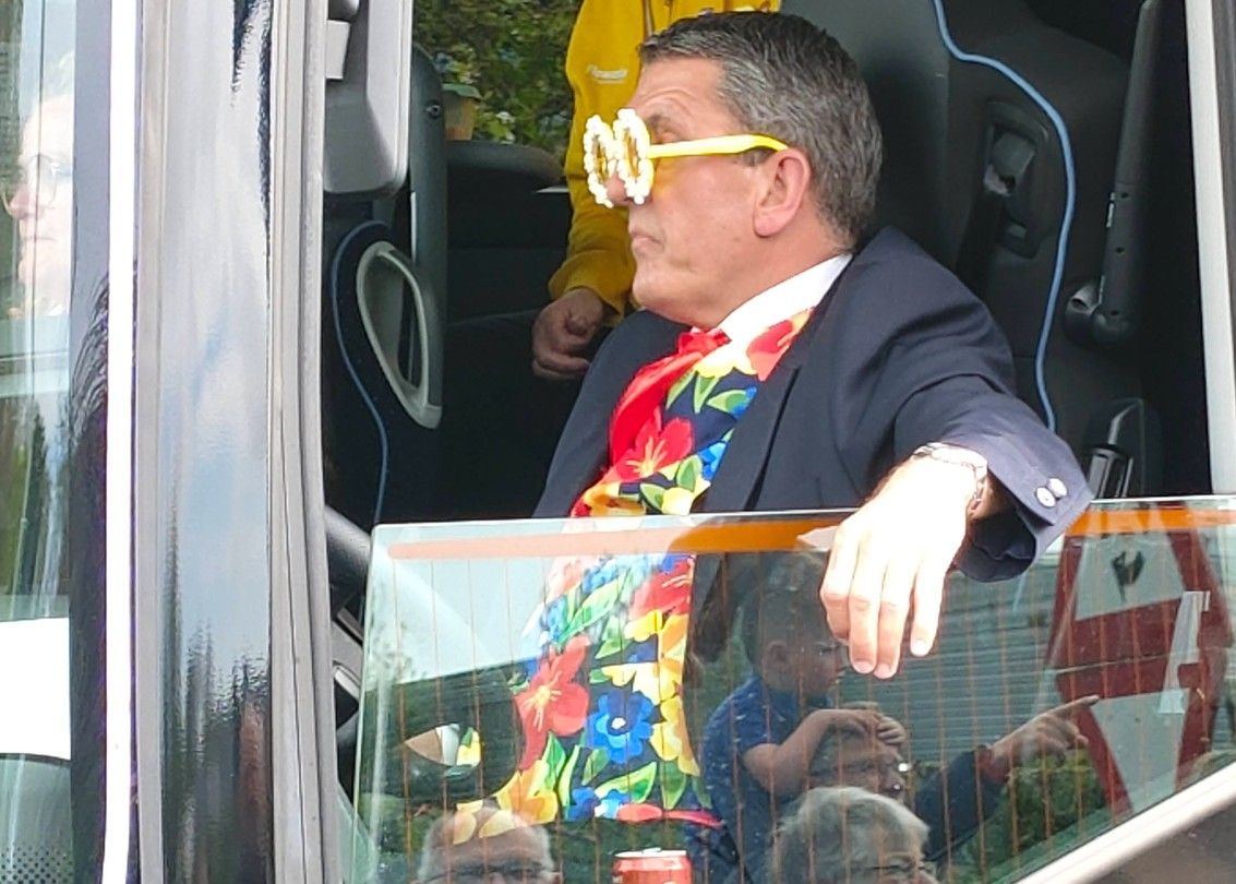 Een man met een opvallende gele bril en een kleurrijk shirt staat in een bus of tram en leunt tegen de leuning.