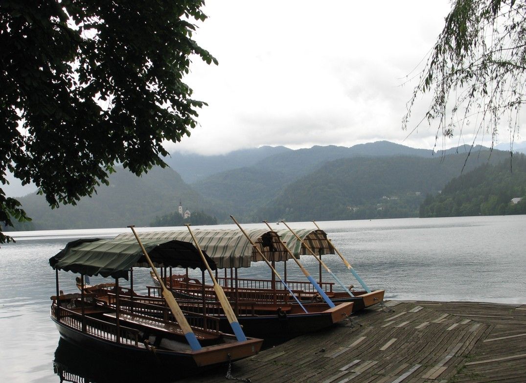 Boten met rieten daken liggen aangemeerd aan de oever van een meer. In de verte zijn bergen te zien onder een bewolkte hemel.