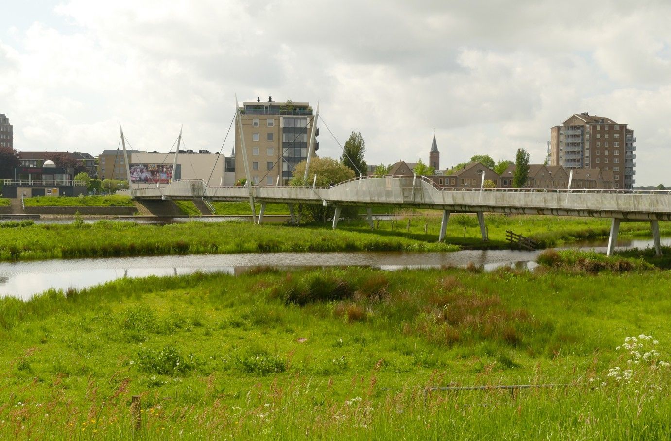 Een voetbrug over een rivier met grasrijke oevers, stadsgebouwen op de achtergrond onder een bewolkte hemel.