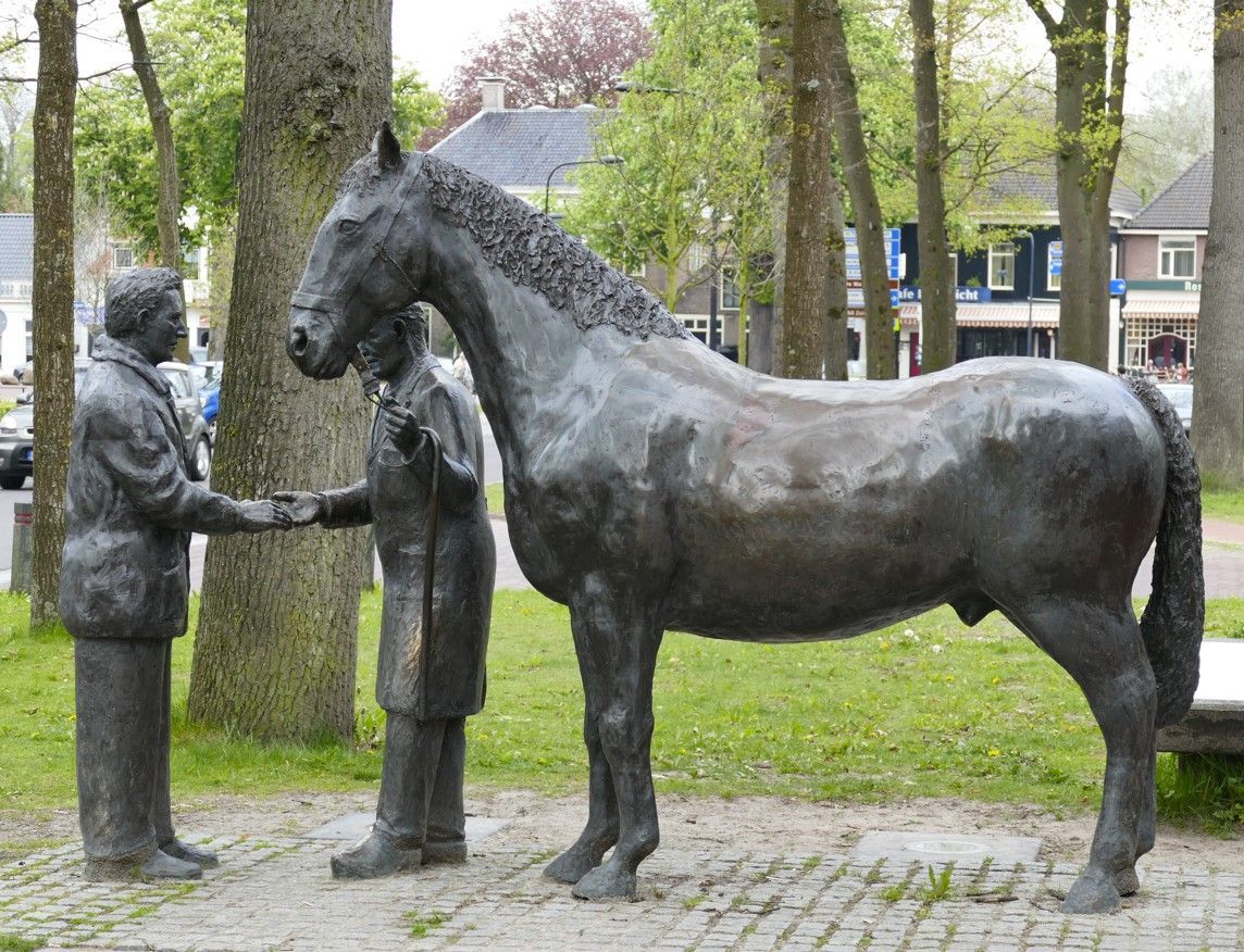Bronzen sculptuur van een paard en twee mannen die elkaar de hand schudden, in de buitenlucht, vlakbij bomen en gebouwen.