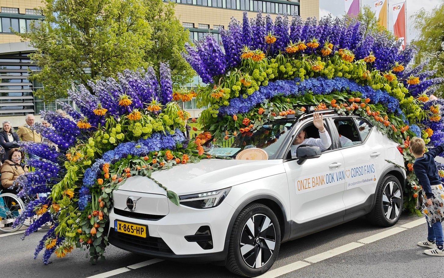 Witte SUV in een bloemenparade met een grote paarse, oranje en blauwe bloemenboog erboven.