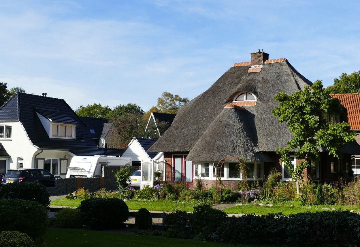 Op een zonnige dag staat een traditioneel huis met een rieten dak naast een modern wit huis, met bomen op de voorgrond.